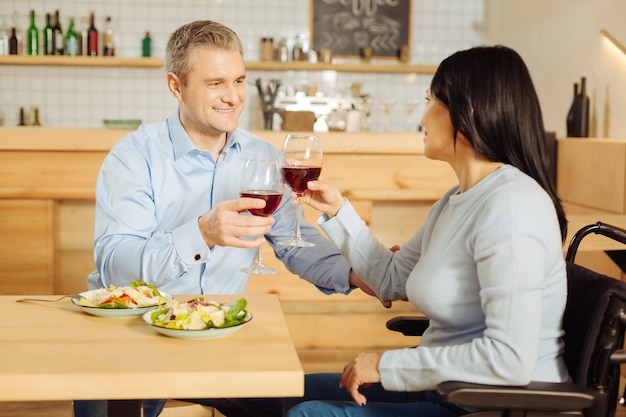 Bel homme blond joyeux souriant et une femme aux cheveux noirs handicapée inspirée souriant et buvant du vin et en train de dîner