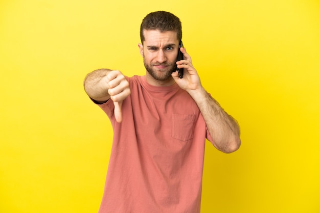 Bel homme blond utilisant un téléphone portable sur fond isolé montrant le pouce vers le bas avec une expression négative