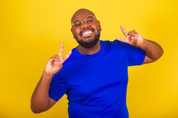 Bel homme brésilien afro portant des lunettes chemise bleue sur fond jaune souriant avec signe de paix et d'amour sur les doigts bonheur joie paix amour