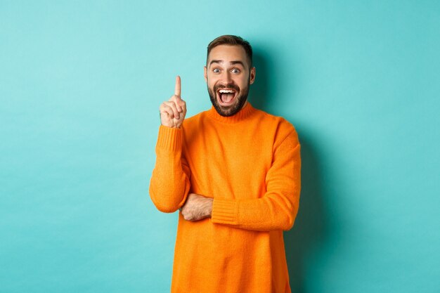 Bel homme caucasien ayant une idée, levant le doigt et disant son plan, debout excité sur fond turquoise.