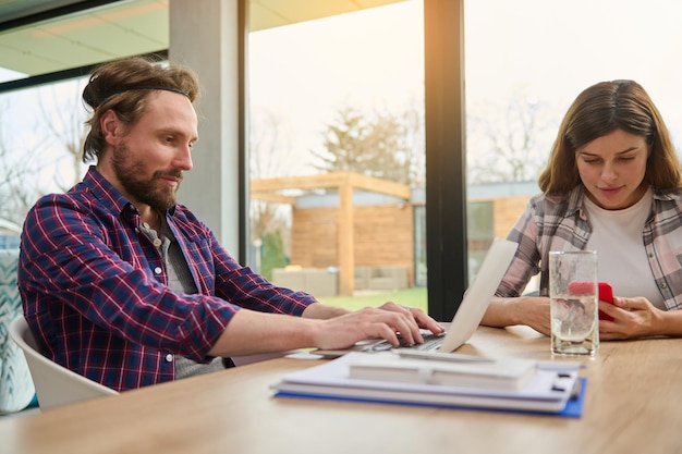 Bel homme caucasien entrepreneur développeur indépendant travaillant à distance sur un ordinateur portable assis à une table à côté de sa femme qui utilise un téléphone portable dans la maison contre des fenêtres donnant sur un jardin