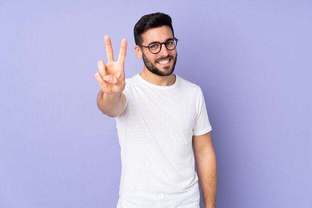 Bel homme caucasien sur mur isolé souriant et montrant le signe de la victoire