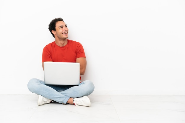 Bel homme caucasien avec un ordinateur portable assis sur le sol heureux et souriant