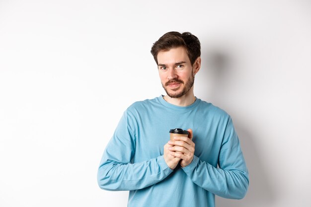 Bel homme caucasien réchauffer les mains avec une tasse de café, souriant à la caméra et regardant tendrement, debout sur fond blanc.