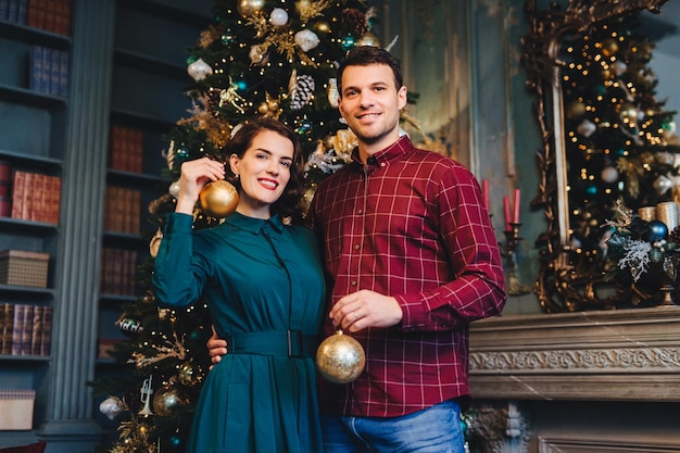 Bel homme avec chaume embrasse sa belle femme tenir des boules de verre dans les mains décorer l'arbre du Nouvel An ensemble préparer les vacances Souriante jeune belle femme et son mari à la maison