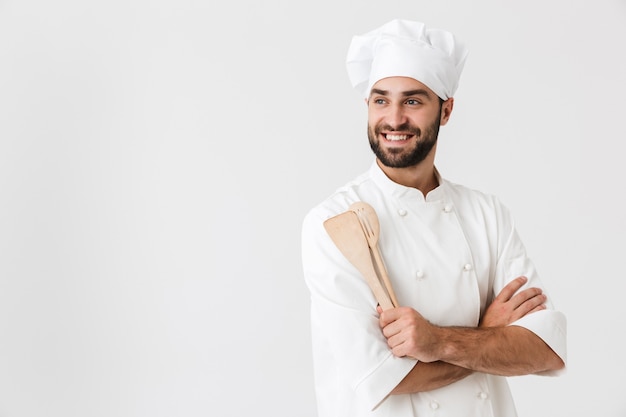 bel homme en chef en uniforme de cuisinier souriant tout en tenant des ustensiles de cuisine en bois isolés sur un mur blanc