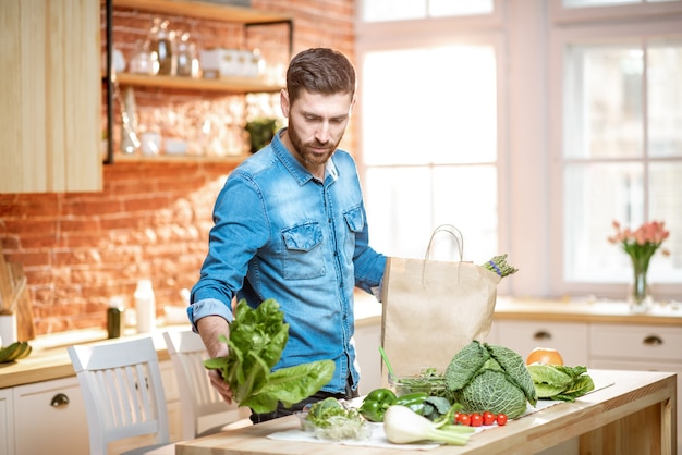 Bel homme en chemise bleue déballant des aliments sains du sac à provisions dans la cuisine