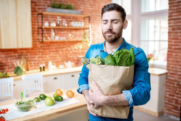 Bel homme en chemise bleue debout avec un sac plein de nourriture verte saine dans la cuisine