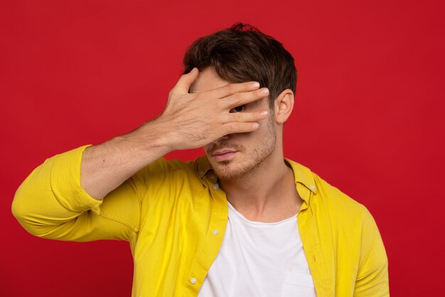 bel homme en chemise jaune couvrant le visage avec la main isolé sur fond rouge