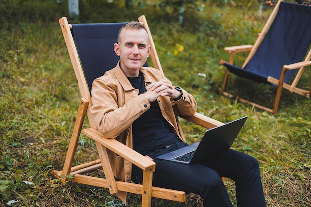 Un bel homme en chemise marron est assis sur une chaise pliante en bois dans le jardin et travaille avec un ordinateur portable Meubles écologiques en bois Travail à distance