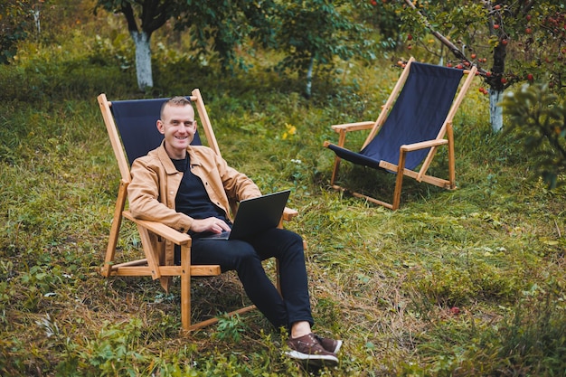 Un bel homme en chemise marron est assis sur une chaise pliante en bois dans le jardin et travaille avec un ordinateur portable Meubles écologiques en bois Travail à distance