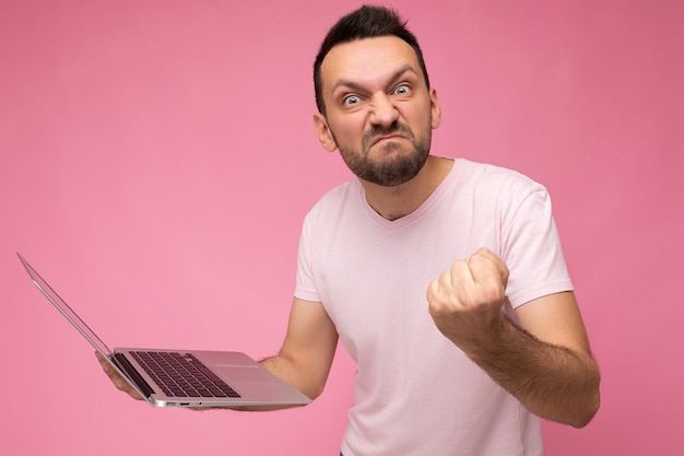 Bel homme en colère tenant un ordinateur portable montrant le poing regardant la caméra en t-shirt sur rose isolé