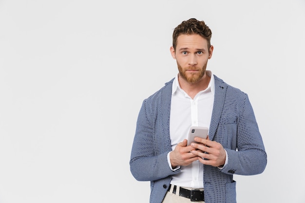 Bel homme confiant portant une veste en tapant sur un smartphone isolé sur un mur blanc