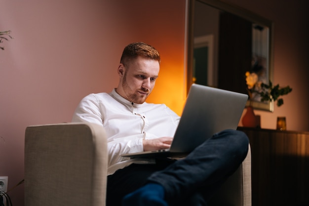 Bel homme confiant utilisant un ordinateur portable assis dans un fauteuil moelleux à la maison dans un salon confortable. Concept de travail à distance à domicile pendant la pandémie de coronavirus COVID-19.