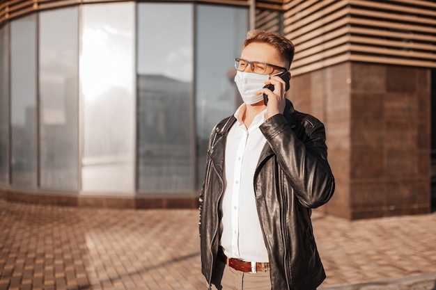 Bel homme dans un masque de protection sur le visage avec des lunettes avec un smartphone dans la rue d'une grande ville. Homme d'affaires parlant au téléphone sur fond urbain