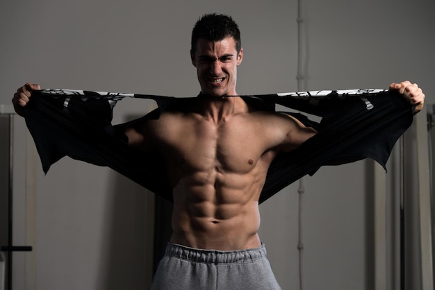 Bel homme debout fort dans la salle de gym et déchirant le t-shirt du modèle de forme physique de bodybuilder athlétique musclé posant après les exercices