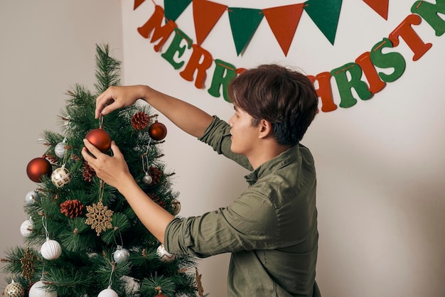 Bel homme décorant le sapin de noël avec des boules et regardant la caméra à la maison