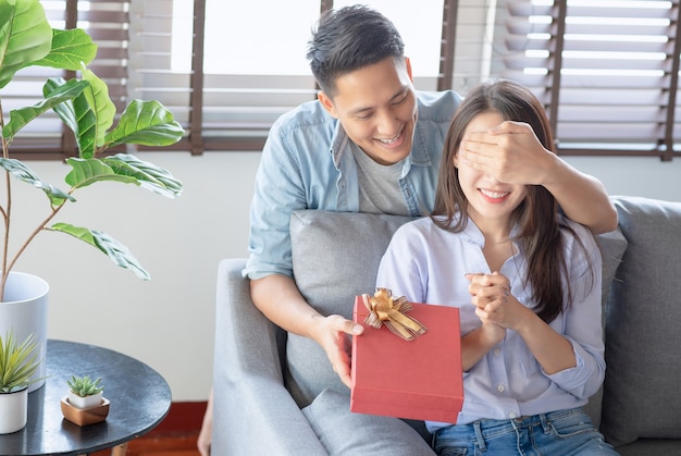 Bel homme donne à sa petite amie une boîte cadeau rouge pour anniversaire surprenant dans le salon à la maison