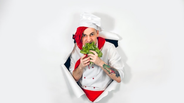 Un bel homme avec des dreadlocks rouges mange des feuilles de laitue Un cuisinier fou en tablier blanc et une cravate rouge mange des ingrédients pour un plat végétarien regardant à travers un fond de papier blanc déchiré en studio