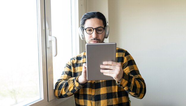 Bel homme avec des écouteurs et des lunettes discutant avec une tablette près d'une fenêtre