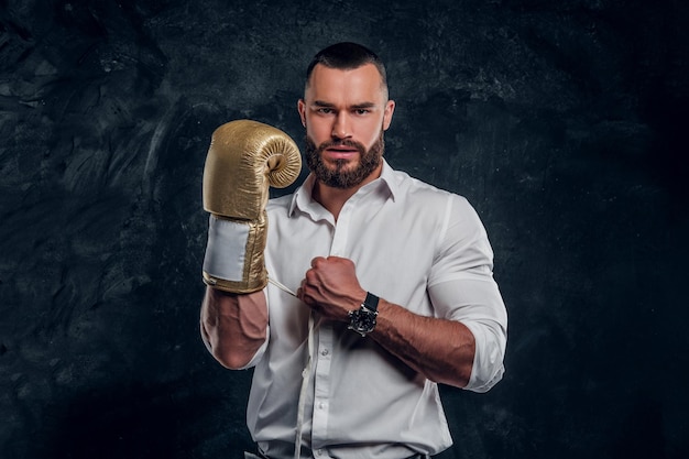 Un bel homme effronté en chemise blanche et avec un gant de boxe doré se tient dans un studio sombre.