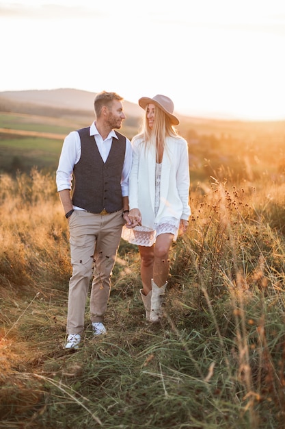 Bel homme élégant en costume rustique et jolie femme bohème en robe, veste, chapeau et bottes de cowboy, marchant sur le terrain