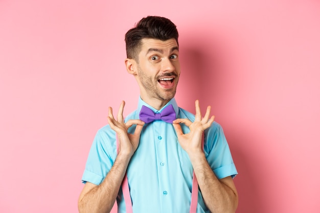 Bel homme élégant fixant un nœud papillon sur le cou et souriant, se préparant pour une date ou une fête, debout sur fond rose.