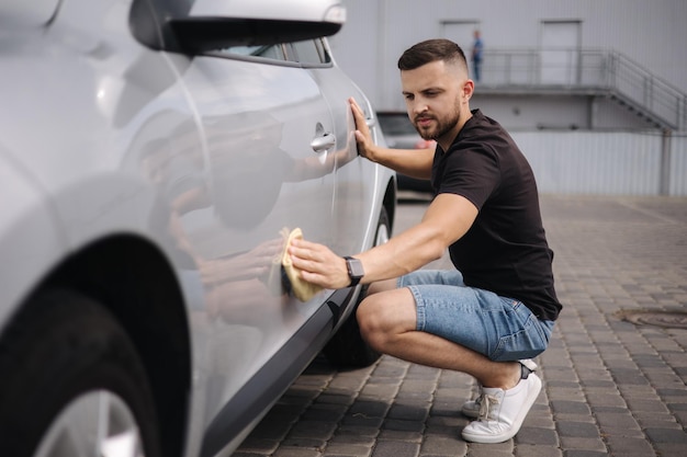 Bel homme essuie une voiture avec un chiffon dans une salle d'exposition dans un lave-auto en libre-service voiture grise voiture polie