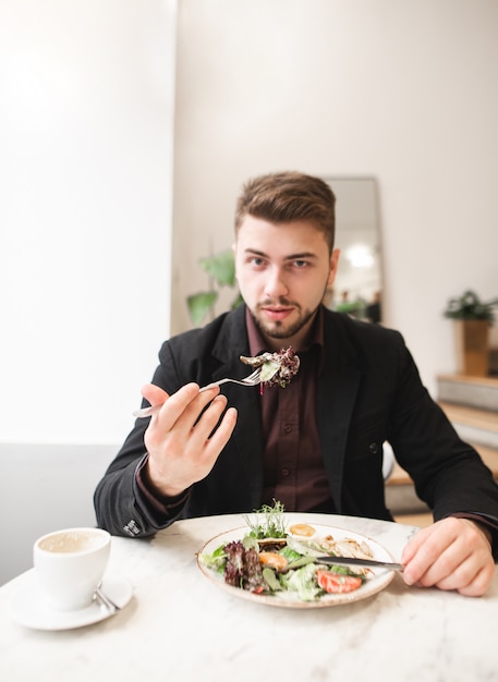 Bel homme est assis dans un restaurant lumineux confortable et mange une assiette de salade et boit du café.