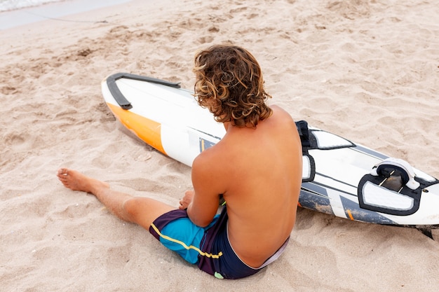 Bel homme est assis sur la plage avec une planche de surf vierge blanche attendre la vague de surf spot en mer océan