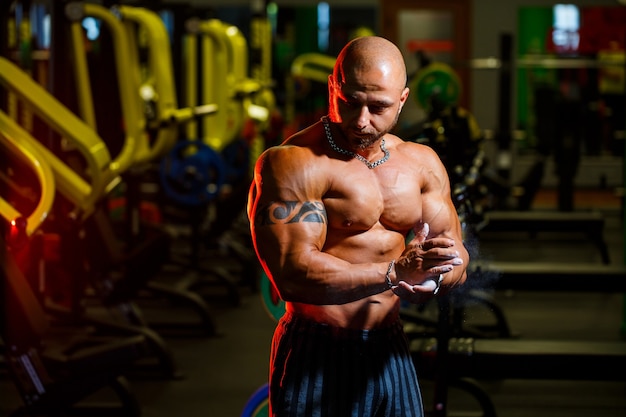 Bel homme fort sportif posant sur le fond de la salle de gym. Un bodybuilder fort avec des abdominaux, des épaules, des biceps, des triceps et une poitrine parfaits.