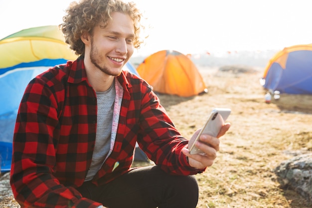 Bel homme gai campant à l'extérieur, utilisant un téléphone portable