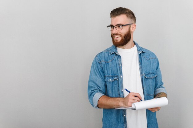 bel homme gai à lunettes écrivant et tenant un presse-papiers isolé sur un mur gris