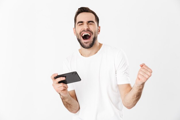 Bel Homme Gai Portant Un T-shirt Vierge Debout Isolé Sur Un Mur Blanc, Jouant à Des Jeux Sur Téléphone Portable