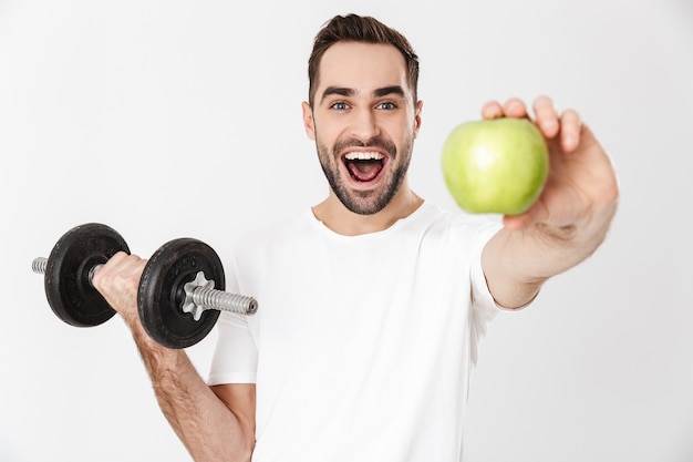 Bel homme gai portant un t-shirt vierge debout isolé sur un mur blanc, montrant une pomme verte, faisant de l'exercice avec un haltère