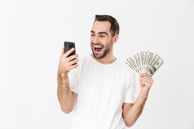 Bel homme gai portant un t-shirt vierge debout isolé sur un mur blanc, utilisant un téléphone portable, montrant des billets en argent