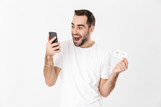 Bel homme gai portant un t-shirt vierge debout isolé sur un mur blanc, utilisant un téléphone portable, montrant une carte de crédit en plastique