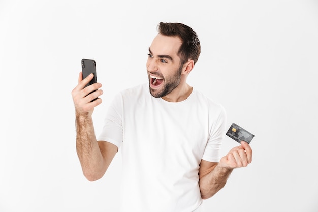 Bel homme gai portant un t-shirt vierge debout isolé sur un mur blanc, utilisant un téléphone portable, montrant une carte de crédit en plastique