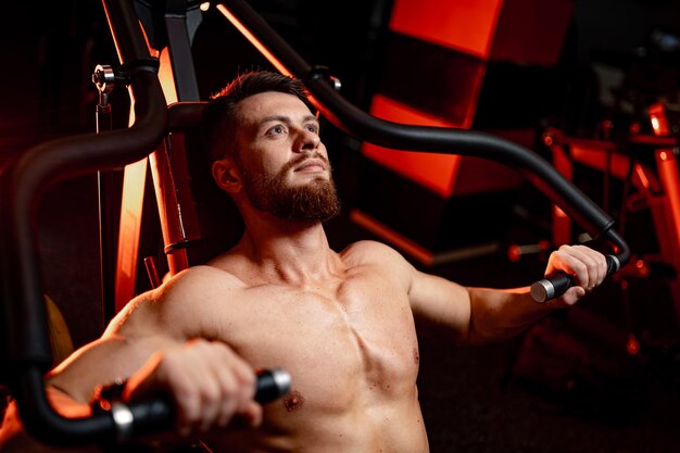 Bel homme avec de gros muscles posant devant la caméra dans la salle de gym Couché sur un banc et poussant des poids Gros plan