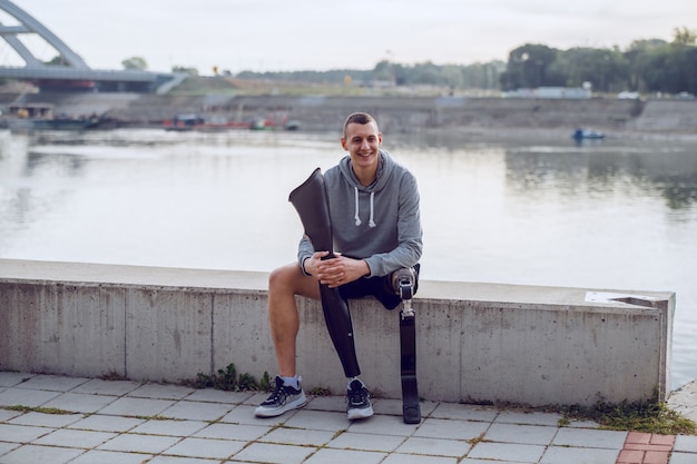 Bel homme handicapé caucasien sportif en vêtements de sport et avec une jambe artificielle assis sur le quai et tenant une autre jambe artificielle.
