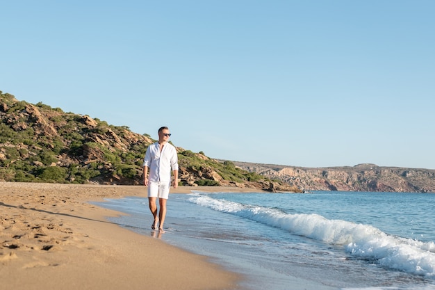 Bel homme heureux en chemise blanche marchant sur la plage