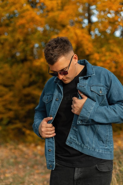 Bel homme hipster élégant avec une coiffure et des lunettes de soleil vintage dans une chemise en jean à la mode et un t-shirt noir marchant dans le parc avec un feuillage d'automne jaune