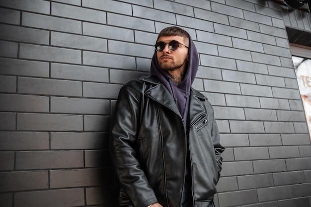 Un bel homme hipster à la mode avec des lunettes de soleil rondes à la mode portant une veste en cuir à la mode et un sweat à capuche se tient près d'un mur de briques noires dans la rue