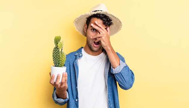 Bel homme hispanique à l'air choqué, effrayé ou terrifié, couvrant le visage avec un agriculteur à la main et un concept de cactus