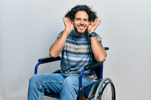 Bel homme hispanique assis sur un fauteuil roulant essayant d'entendre les deux mains sur le geste de l'oreille, curieux de potins. problème d'audition, sourd