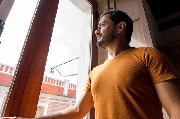 Un bel homme hispanique barbu avec un t-shirt orange regardant par la fenêtre