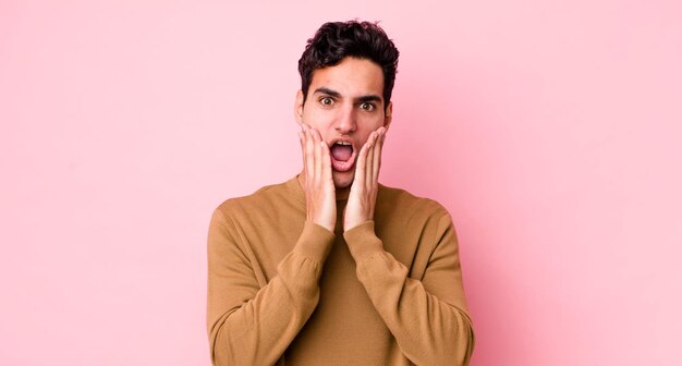 Bel homme hispanique se sentant choqué et effrayé, terrifié avec la bouche ouverte et les mains sur les joues