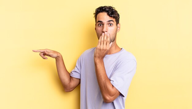 Bel homme hispanique se sentant heureux, couvrant la bouche avec la main et pointant vers l'espace de copie latéral