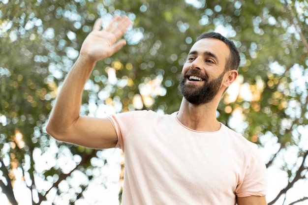 Bel homme hispanique souriant, agitant la main, regardant loin, debout dans le parc. Mouvement, mise au point sélective