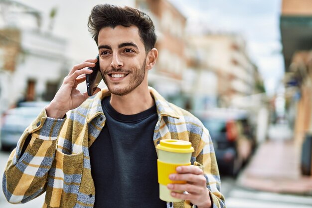 Bel homme hispanique souriant heureux et confiant à la ville parlant au téléphone et buvant une tasse de café